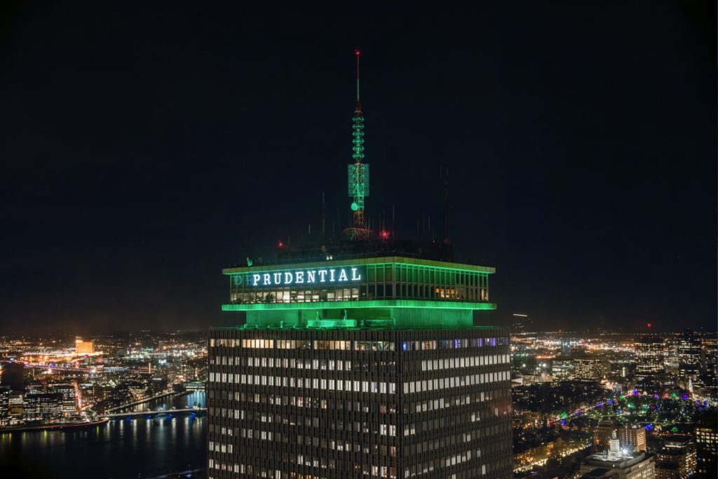 View of the top if the Prudential Center building lit in green lights. The Boston city skyline at night is visible in the background. 